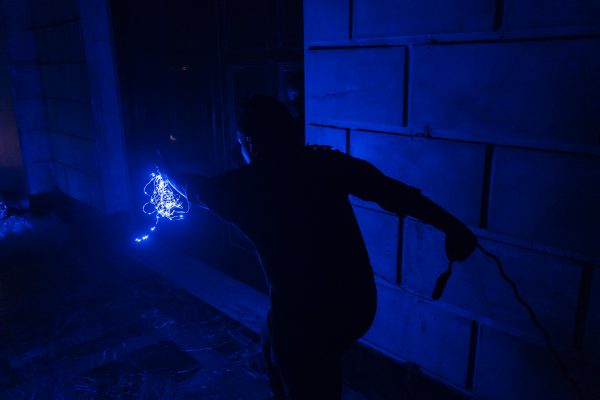alt text: Photo of Shantelle Courvoisier lunging forward in the dark ISSUE Project Room space, holding a blue string light in her left hand and a wire in her right hand.
